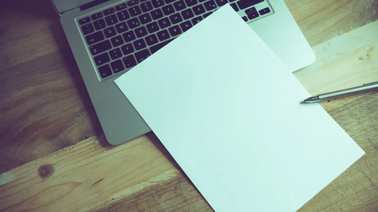 Folha de papel em branco e caneta sobre um notebook aberto, em mesa de madeira.