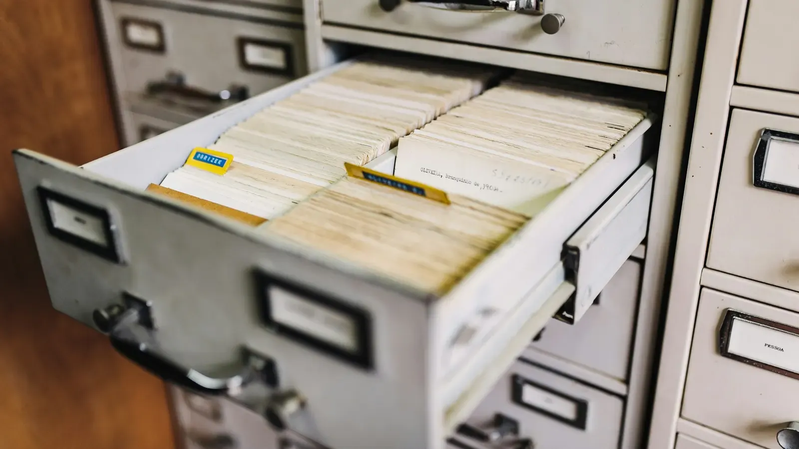 Gaveta de arquivo de aço aberta, cheia de fichas de papel com nomes escritos à mão.