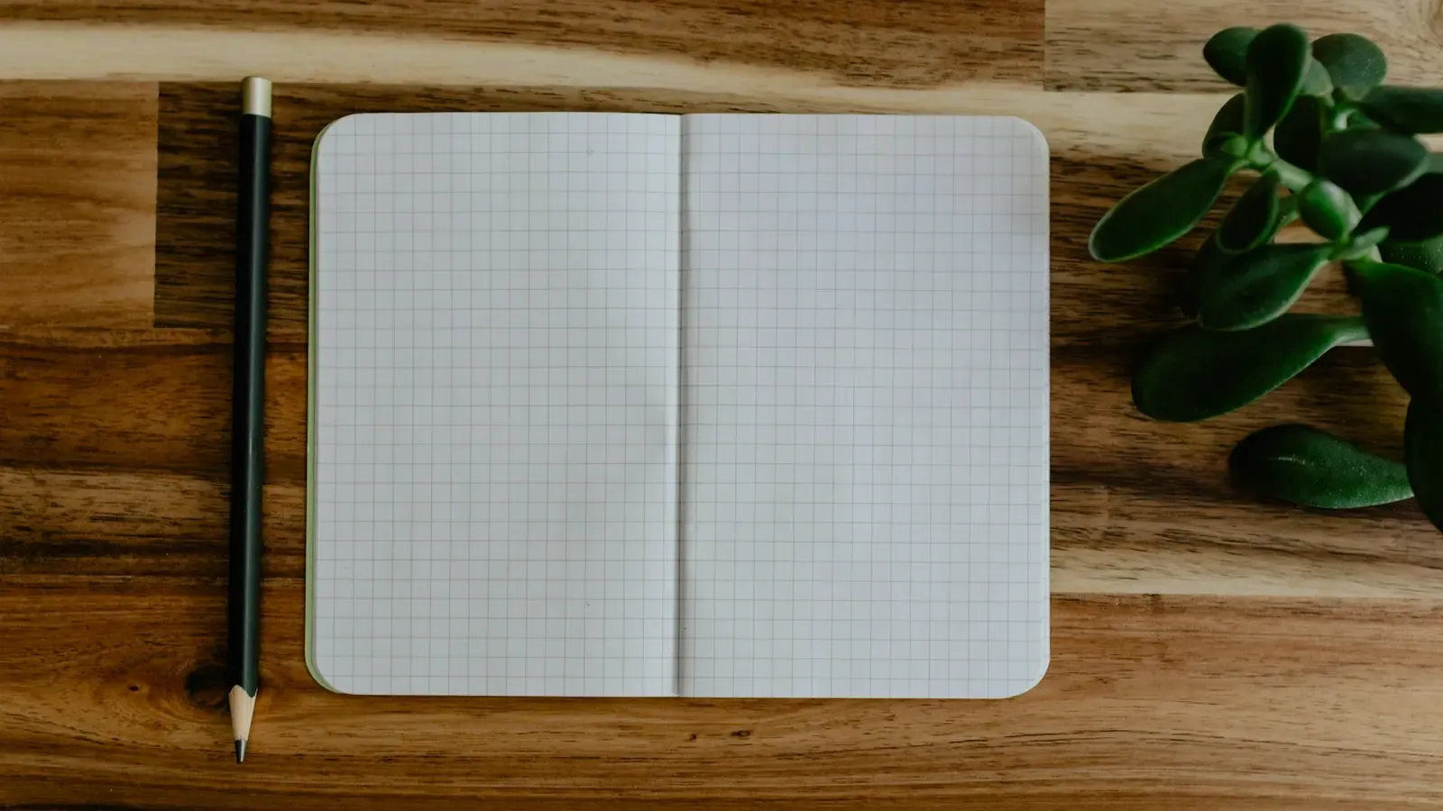 Caderno quadriculado aberto em página em branco, ao lado de um lápis, sobre mesa de madeira.